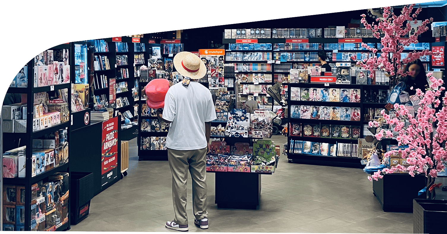 Innenansicht des Manga Cult Stores mit gefüllten Regalen voller Manga, Figuren und Merchandise. Im Zentrum steht ein Besucher mit Strohhut und Plüschfigur in der Hand. Rechts blüht ein dekorativer Kirschblütenbaum. Sichtbare Bereiche für Neuheiten, Merchandising und Manga Cult-Titel.