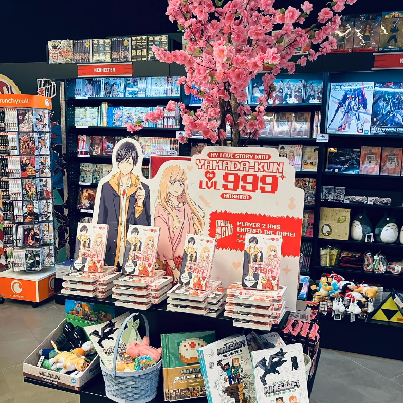 Buchdisplay im Manga Cult Store für „My Love Story with Yamada-kun at Lv999“ von Mashiro: Aufsteller mit lebensgroßen Charakterillustrationen, mehreren gestapelten Manga-Bänden und Deko-Kirschblüten. Im Hintergrund Regale mit Neuheiten und Merchandise.