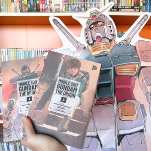 Eine Hand hält die Bände 1 und 2 von "Mobile Suit Gundam – Origin" vor einem großen Gundam-Aufsteller, mit einem Manga-Regal im Hintergrund. 