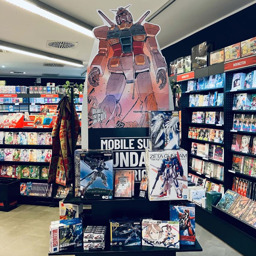 Hintrgrund: Schwarze Bücherregale mit Mangas. Vordergrund: Tisch mit großem Mobile-Suit-Gundam-Aufsteller und Gundam Mangas, Gunplas und Figuren