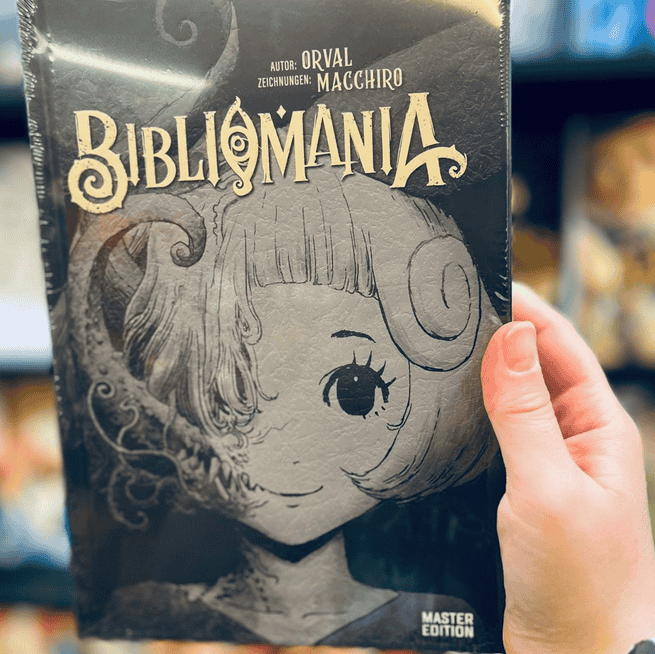 Eine Hand zeigt im Vordergrund die Master-Edition der Manga Serie Bibliomania von Orval. Im Hintergrund sieht man den schwarzen Bücherregal voller Bücher vom Manga Cult Store