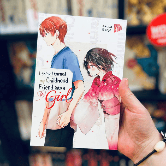 Eine Hand zeigt im Vordergrund Band 4 der Manga Serie I Think I Turned My Childhoodfriend Into A Girl von Azusa Banjo. Im Hintergrund sieht man den schwarzen Bücherregal voller Bücher vom Manga Cult Store