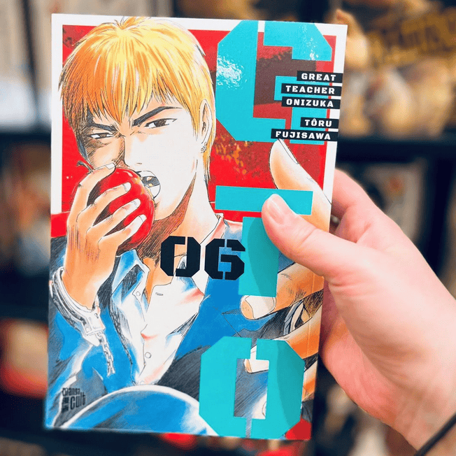 Eine Hand zeigt im Vordergrund Band 6 der Manga Serie GTO von Toru Fujisawa. Im Hintergrund sieht man den schwarzen Bücherregal voller Bücher vom Manga Cult Store