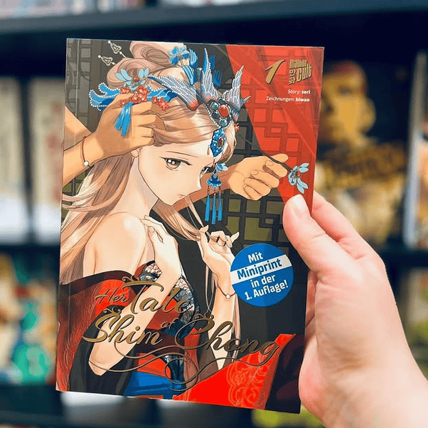 Eine Hand zeigt im Vordergrund Band 1 der Manhwa-Serie Her Tale of Shim Chong von Seri und Biwan. Im Hintergrund sieht man den schwarzen Bücherregal voller Bücher vom Manga Cult Store