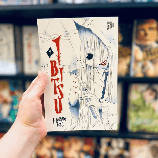 Eine Hand zeigt im Vordergrund Band 1 der Manga Serie Ibitsu von Ryo Haruto. Im Hintergrund sieht man den schwarzen Bücherregal voller Bücher vom Manga Cult Store