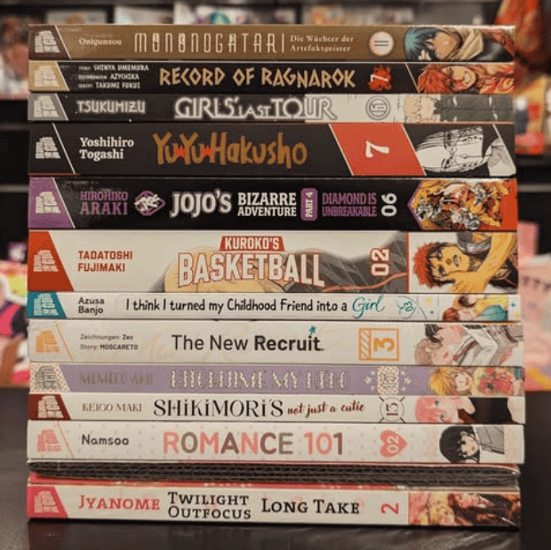 Ein Stapel Manga-Bücher liegt auf einem Tisch. Sichtbare Titel sind unter anderem „Mononogatari“, „Record of Ragnarok“, „Girls’ Last Tour“, „Yu Yu Hakusho“, „JoJo’s Bizarre Adventure: Diamond is Unbreakable“, „Kuroko’s Basketball“, „Shikimori’s Not Just a Cutie“ und „Romance 101“. Im Hintergrund stehen unscharfe Figuren als Dekoration.