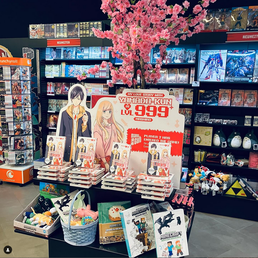 Im Hintergrund: Schwarze Bücherregale voller Manga. im Vordergrund: Verkaufstisch mit Sakura-Baum, einem großen Aufsteller der Manga-Serie My Love Story with Yamada-kun at LVL 999, Manga-Bücher. Unten: Ausmalbücher und Rezptbücher von MINECRAFT