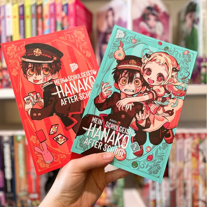Im Hintergrund: Buchregal mit Mangas. Im Vordergrund: Hand hält Band 1 und 2 der Manga-Reihe: Mein Schulgeist Hanako after School