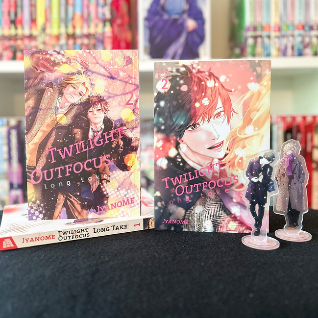 Präsentation von „Twilight Outfocus – Long Take“ Band 2 von Jyanome: Zwei Varianten des Manga-Covers auf schwarzem Tisch, daneben Acryl-Aufsteller mit den Hauptcharakteren. Im Hintergrund ein Regal mit weiteren Manga-Titeln.